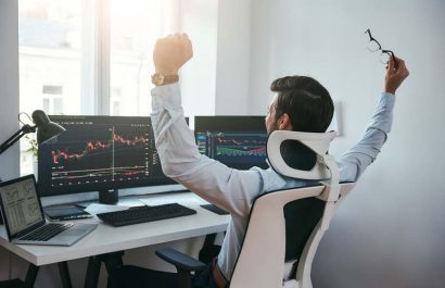 Yes! Back view of stock trader with raised hands looking at multiple computer screens with data and charts and feeling happy while sitting in modern office
