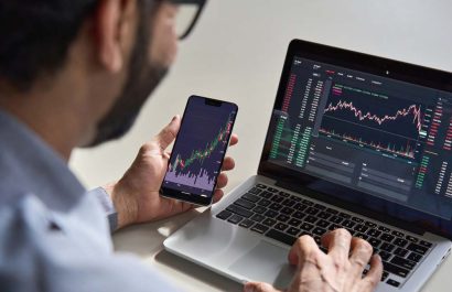 Business man trader investor analyst using mobile phone app analytics for cryptocurrency financial market analysis, trading data index chart graph on smartphone and laptop screen. Over shoulder view
