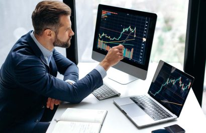 Focused smart successful caucasian male stock investor, broker, sits at his work desk, uses computer and laptop, concentrated analyzes risks and prospects, rise or fall of cryptocurrency coin
